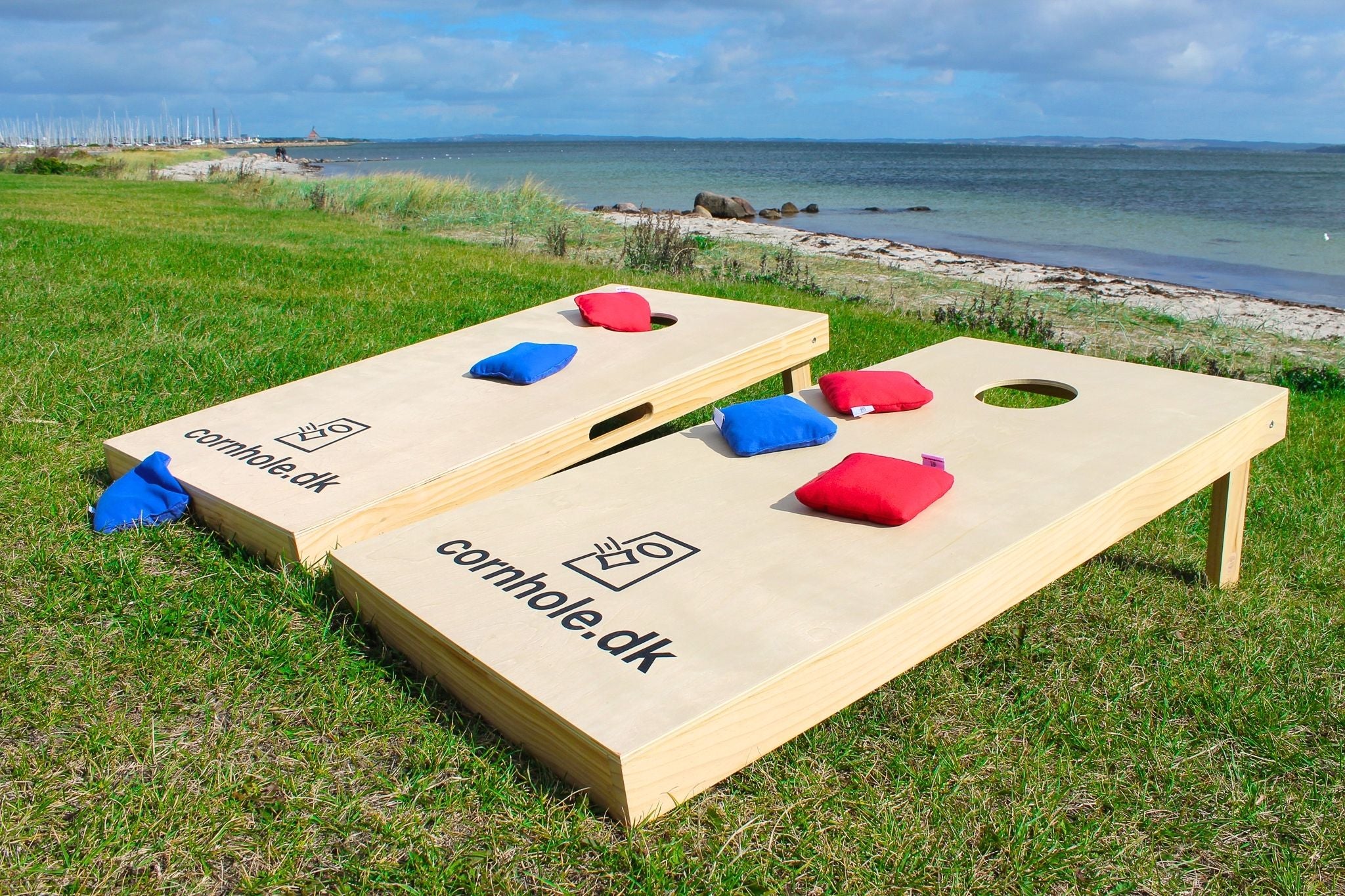 CornholesÊt i de officielle mÄl 120x60 cm fra cornhole.dk stÄr pÄ en grÊsplÊne foran en strand med cornholeposer i farverne rÞd og blÄ fra cornhole.dk