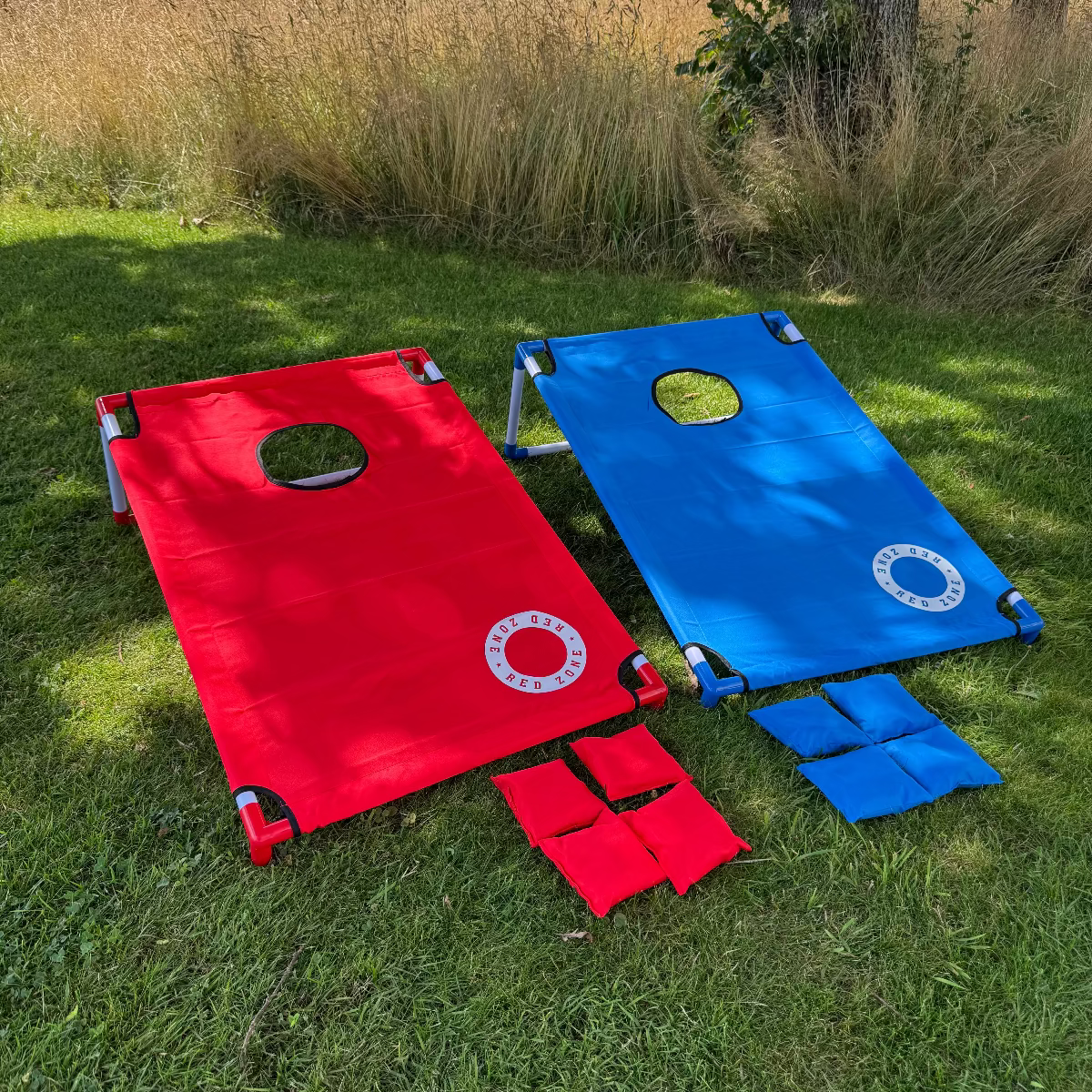 Cornhole strandsÊt 2 brÊt i rÞd og blÄ stÄr pÄ grÊsset med kasteposer i rÞd og blÄ cornholeposer og Êrteposer i et Êrteposekast spil
