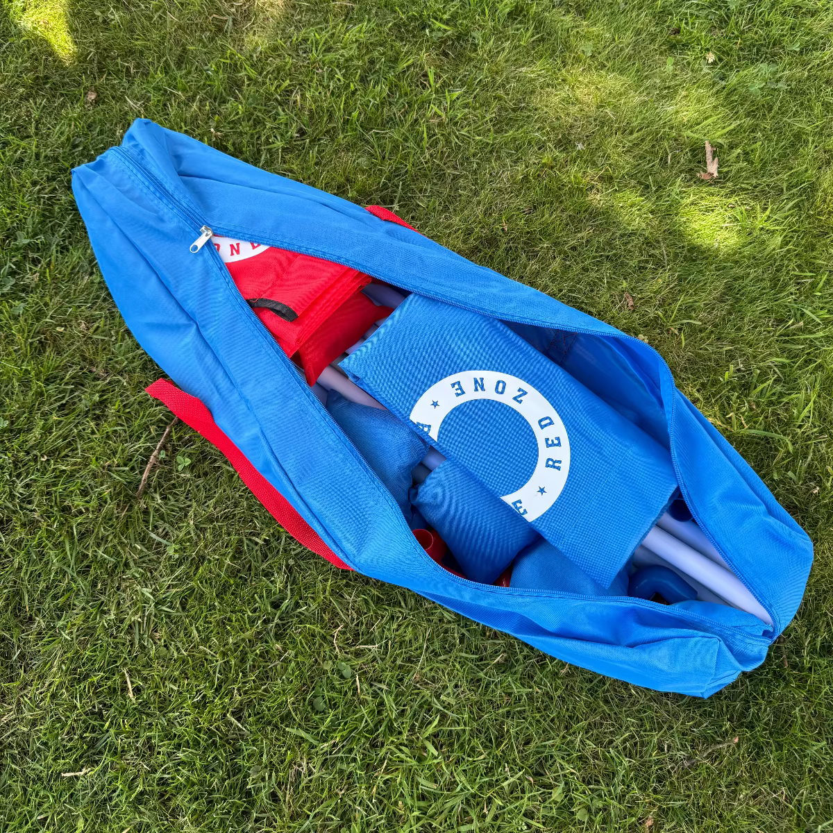Cornhole strand sÊt pakket ned i en let og smart bÊretaske som medfÞlge pÄ grÞnt grÊs. Tasken er blÄ med 2 rÞde cornholebrÊt og 8 rÞde og blÄ cornhole kasteposer, cornholeposer og Êrteposer til et Êrteposekastespil.