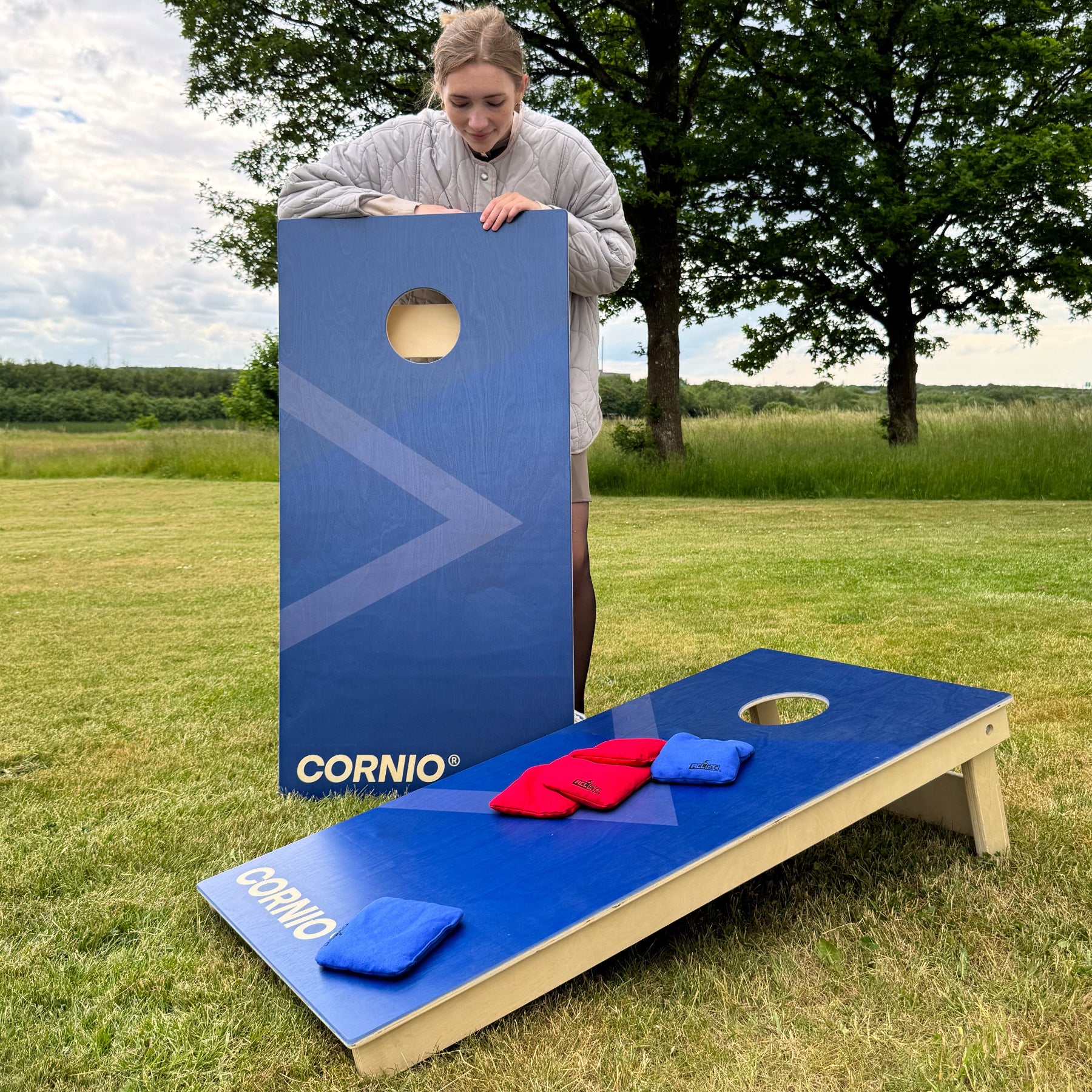 Cornhole game outdoors blue uv printed game with cornio logo and cornholebags in 12mm plywood tops extr asolit with leg brace and cornholebags all weather blue and red, outdoors in grass in the summer.
