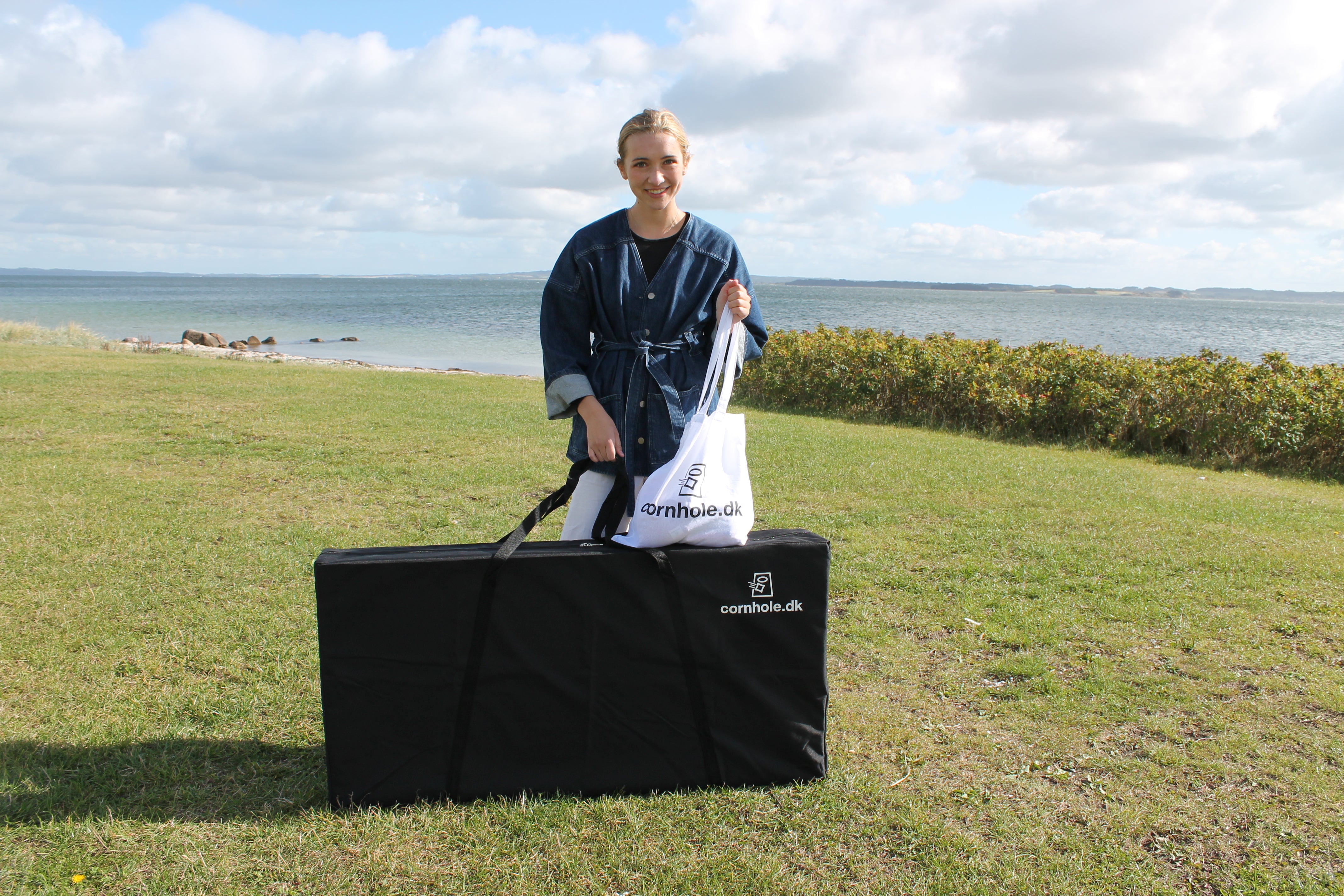 cornhole spil med pige og mulepose med cornhole.dk logo stÄr pÄ en grÊsplÊne ved havet og standen