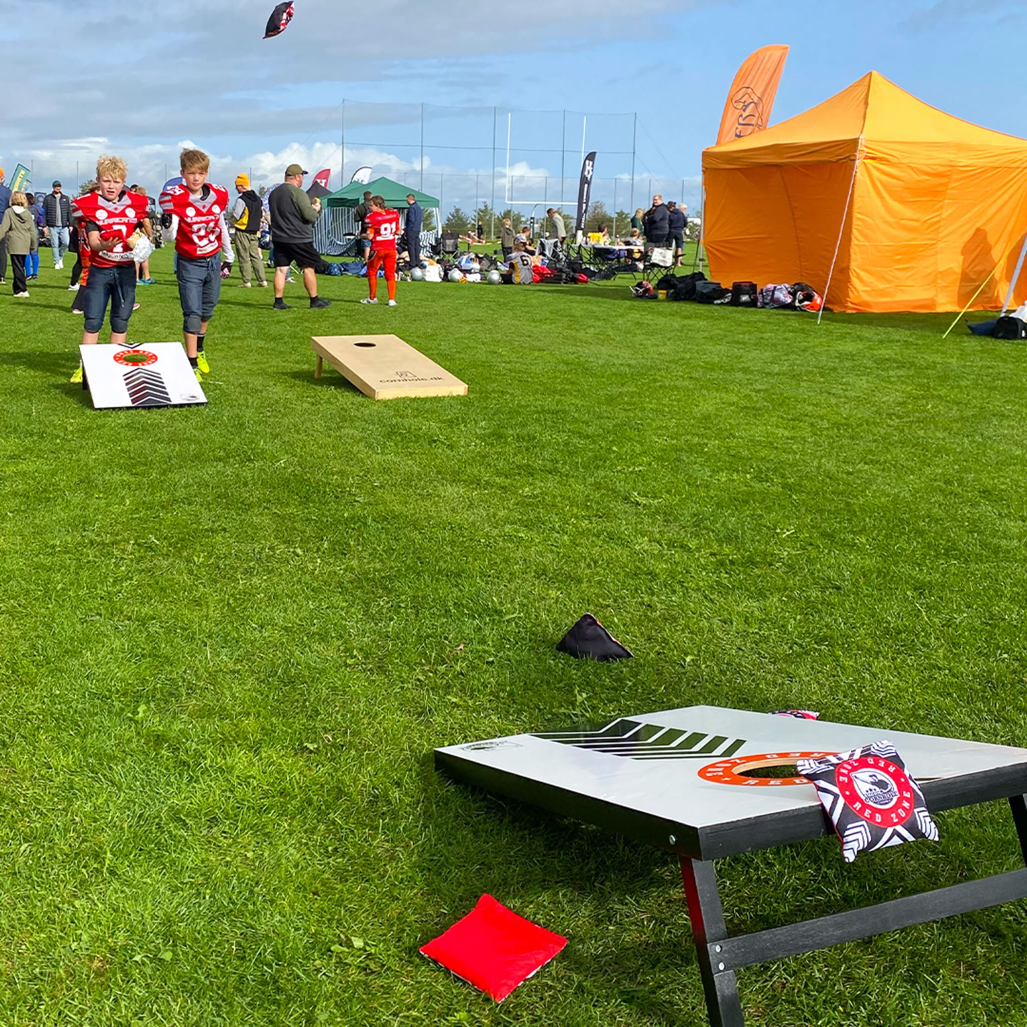BÞrn spiller cornhole med redzone cornholesÊt pÄ en grÊsplÊne ifÞrt udstyr til amerikansk fodbold med et orange telt i baggrunden