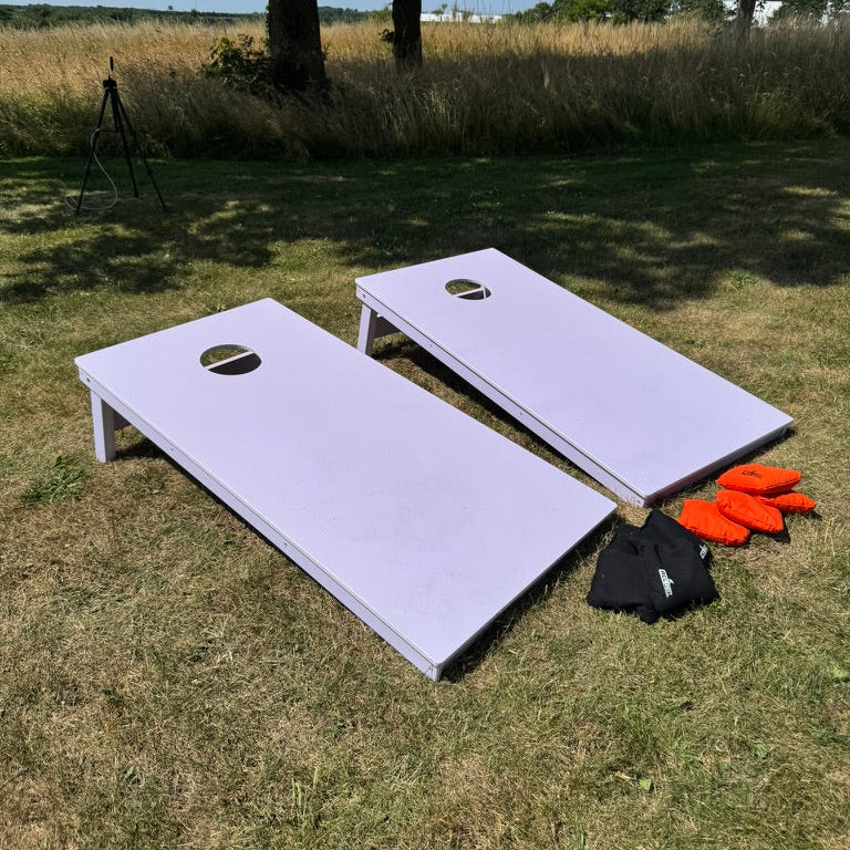 Cornhole spil i pcv vejrbestandigt og 100% vandtæt udendørsspil hvid pvc til sommerhus fester og andet står på en græsplæne med orange og sorte cornhole kasteposer
